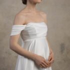 Side-front view of an off-the-shoulder maternity gown with a draped bodice and flowing skirt.